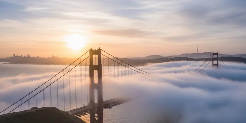 Most Golden Gate wyłaniający się z niskiej mgły o wschodzie słońca, pylony w kolorze International Orange na tle bladego nieba